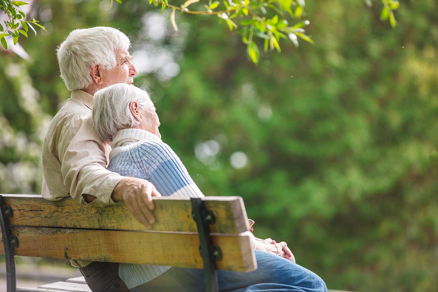 An elderly couple sits on a wooden bench, the woman resting her head on the man's shoulder, enjoying the lush greenery that surrounds them. A moment of serenity made even sweeter with the peace that in-home care provides, allowing them to cherish their time together.